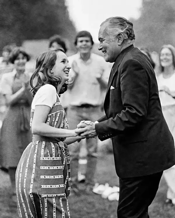 Father Theodore Hesburgh, in a suit, smiles broadly while shaking hands with a young woman wearing a striped apron dress. She smiles back, and a cheering, blurred crowd stands in the background at an outdoor event.