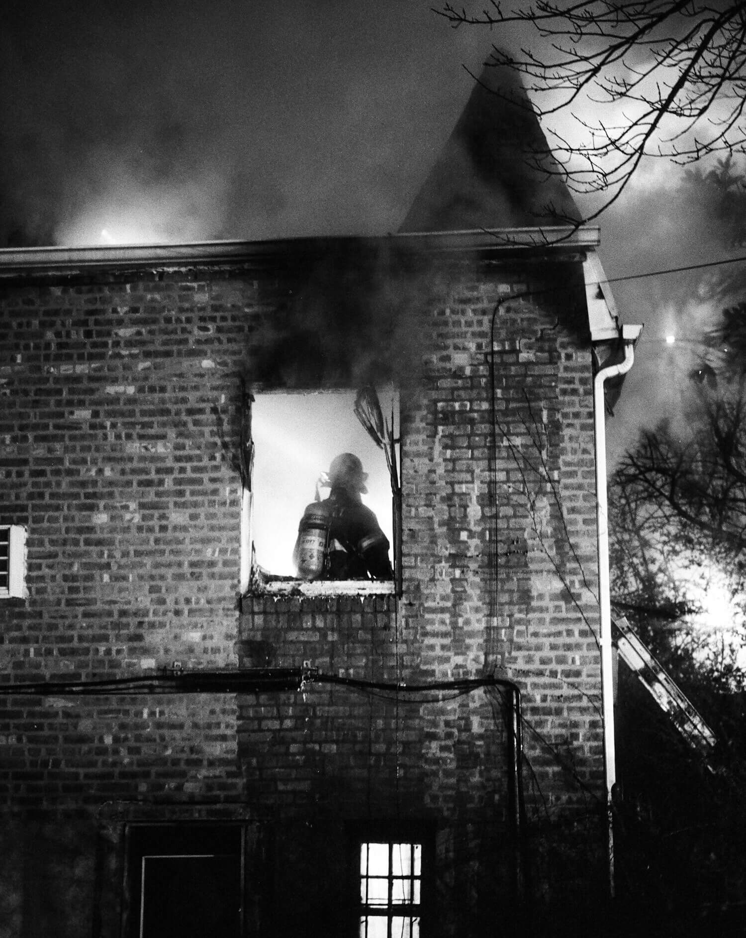 Silhouetted firefighter in helmet and gear stands in a bright, smoky window of a dark brick building at night.