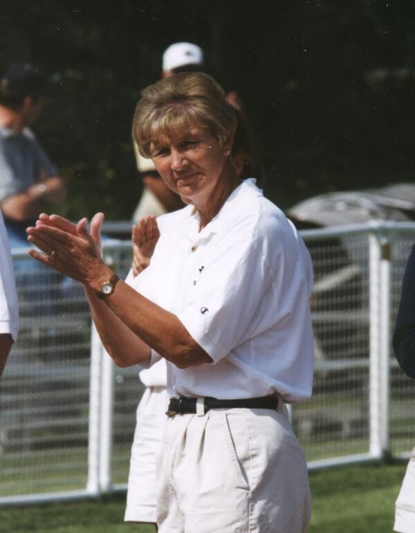 Woman with blonde hair in white polo shirt and tan pleated shorts claps, smiling faintly outdoors.