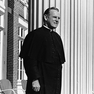 Father Theodore Hesburgh, wearing clerical attire with a cape, smiles outdoors by classical columns.