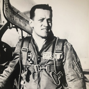 Black and white portrait of Captain Kelly, a pilot with a determined look, in a flight suit and harness in a cockpit.