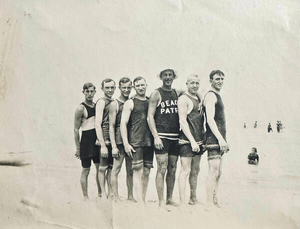 Seven men in vintage dark swimsuits stand barefoot on a sandy beach. One wears a hat and a "BEACH PATR" top, smiling.
