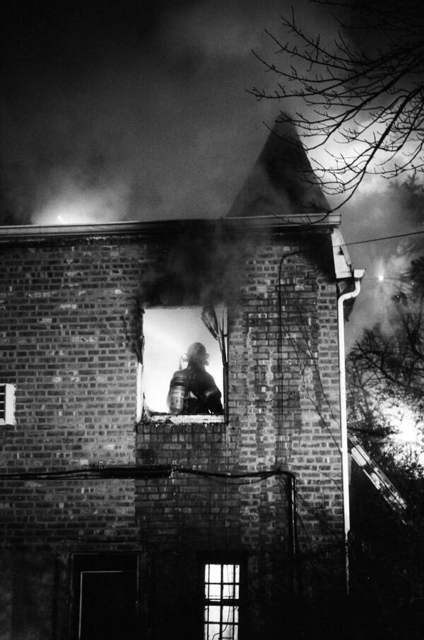 A firefighter, silhouetted against interior light, looks out a smoky window of a brick building at night.