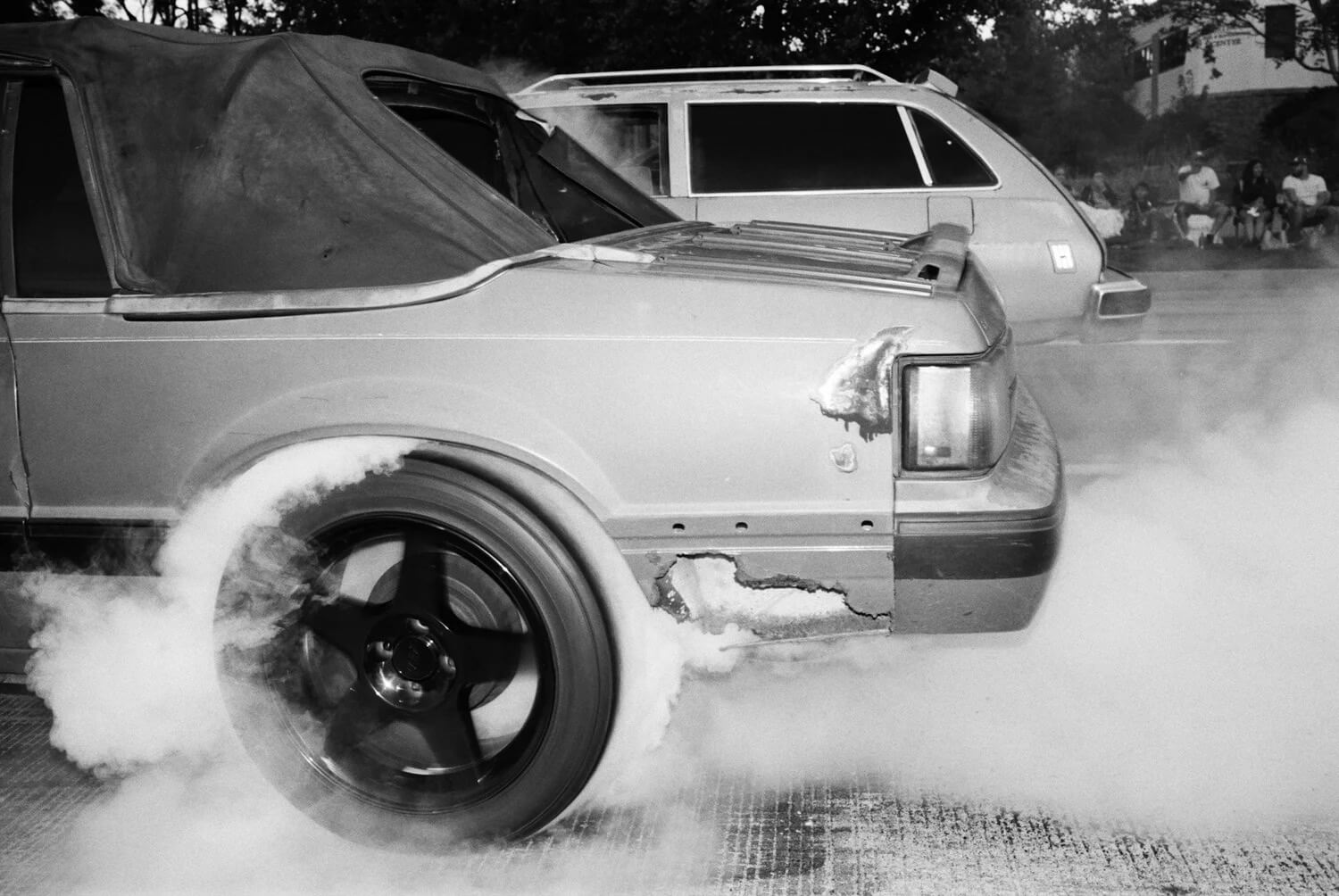 Black and white: A light convertible's rear wheel spins on pavement, producing white smoke from a burnout. Spectators watch.
