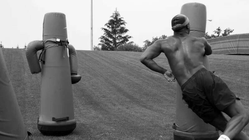 A sweaty, muscular athlete in shorts pushes a tall football tackling dummy on a grassy practice field.