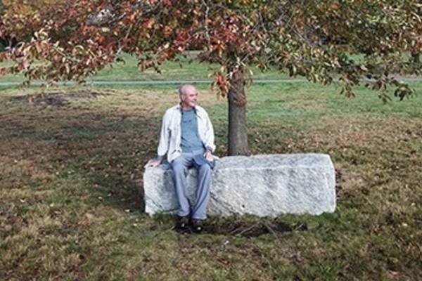 A bald man in a light shirt and blue pants sits on a stone bench under a tree with red autumn leaves, smiling.