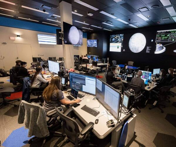 Several people work intently at computers in a dark control room with multiple large screens showing space imagery and data.