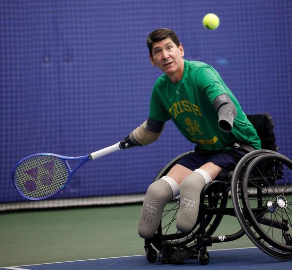 Focused man in green "Irish" shirt in wheelchair swings racket at a tennis ball on indoor court. He has prosthetic limbs.