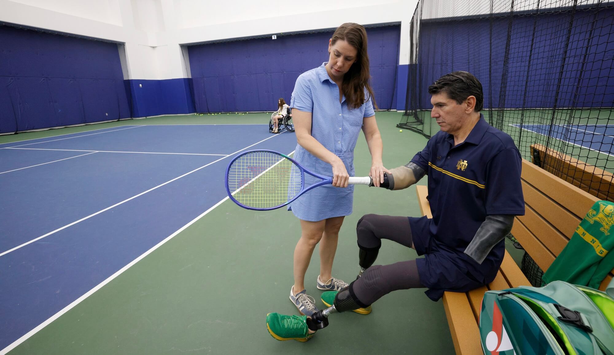 Woman in blue dress assists man in ND polo with prosthetics, connecting a tennis racket to his prosthetic arm.