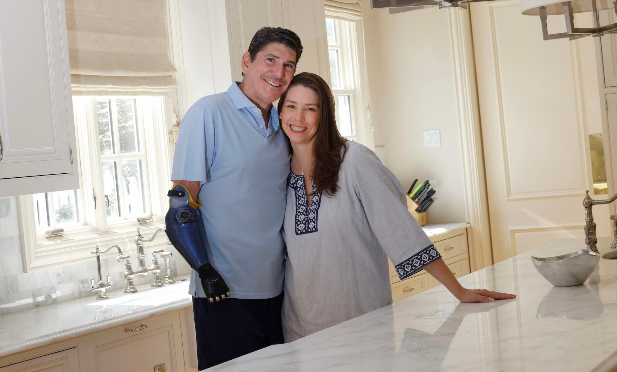 A smiling man in a blue polo shirt with a blue prosthetic arm hugs a woman in a grey embroidered tunic in a bright kitchen.