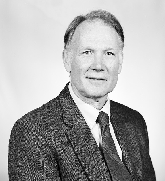 Older man with light hair, smiling gently, wearing a dark herringbone jacket, white shirt, and striped tie.