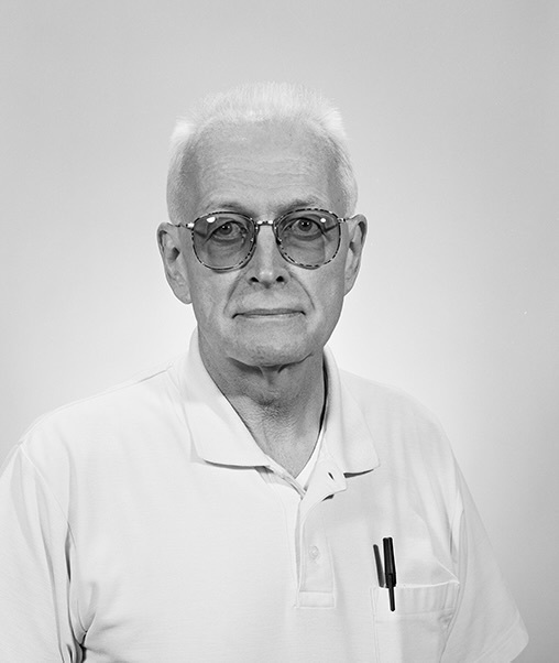 Older man with white hair and tinted glasses, wearing a light polo shirt with a pen in the pocket. Neutral expression.