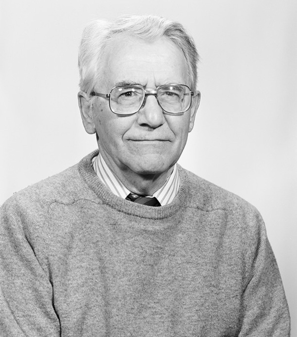 Older man with gray hair, glasses, light-toned striped shirt, dark tie, and light gray sweater. Neutral expression.