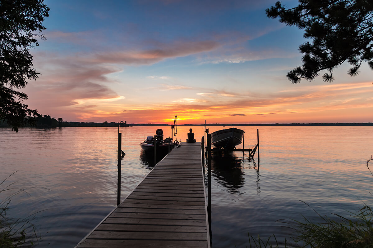 As the sun sets in the distance over a lake, a person in silhouette sits at the end of a pier in between two docked boats.