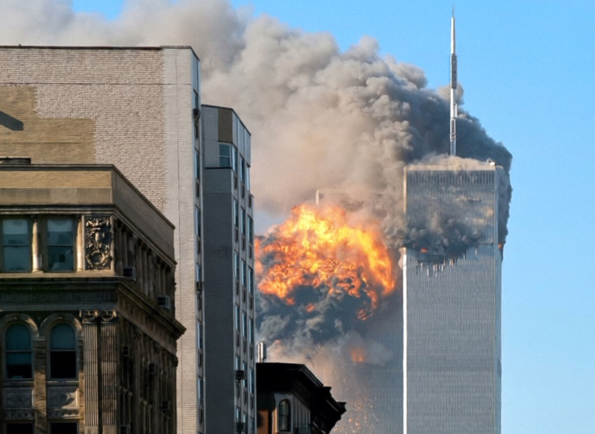 Flames and smoke pour from two skyscapers in the distance with undamaged smaller buildings in the foreground