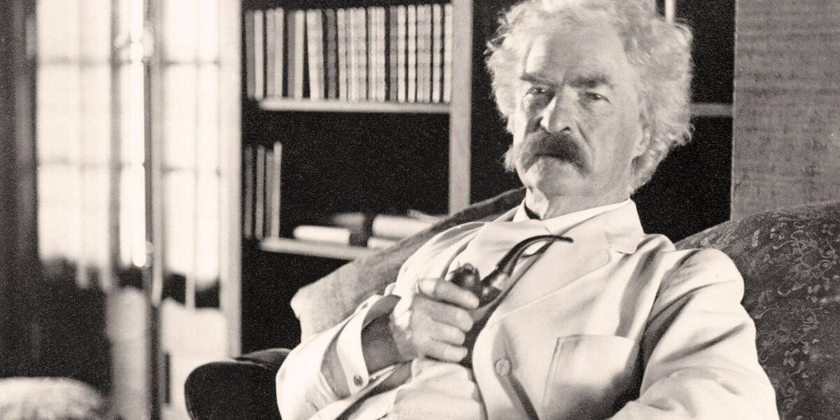 Author Mark Twain, with white hair, a thick moustache and wearing a white suit, leans back in his chair, holding a pipe.
