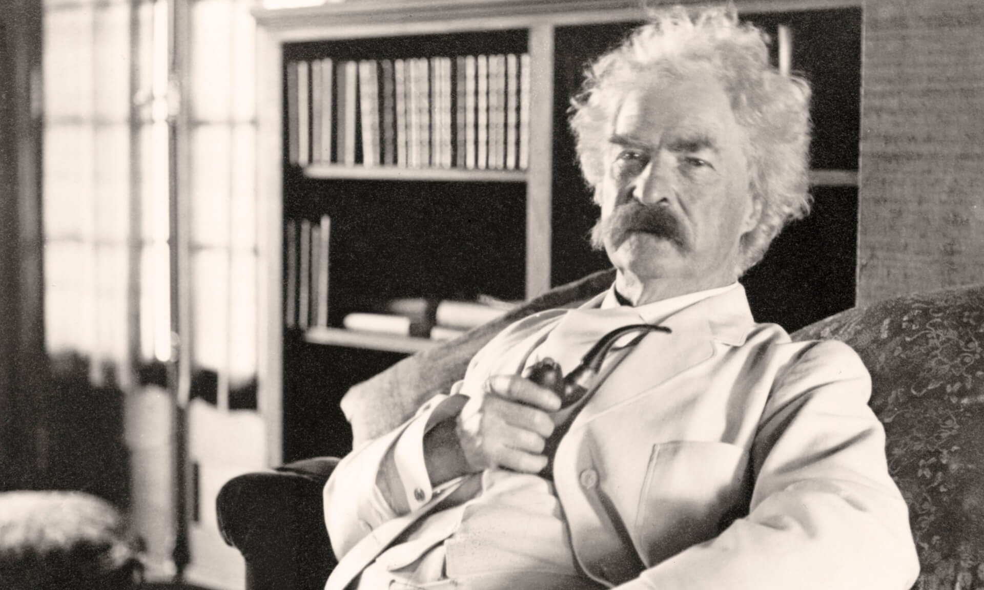 Author Mark Twain, with white hair, a thick moustache and wearing a white suit, leans back in his chair, holding a pipe.
