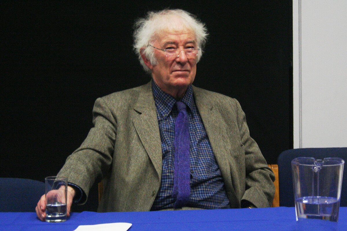Poet Seamus Heaney, with white hair and glasses, sits holding a glass of water, wearing a green jacket over a blue shirt and tie.